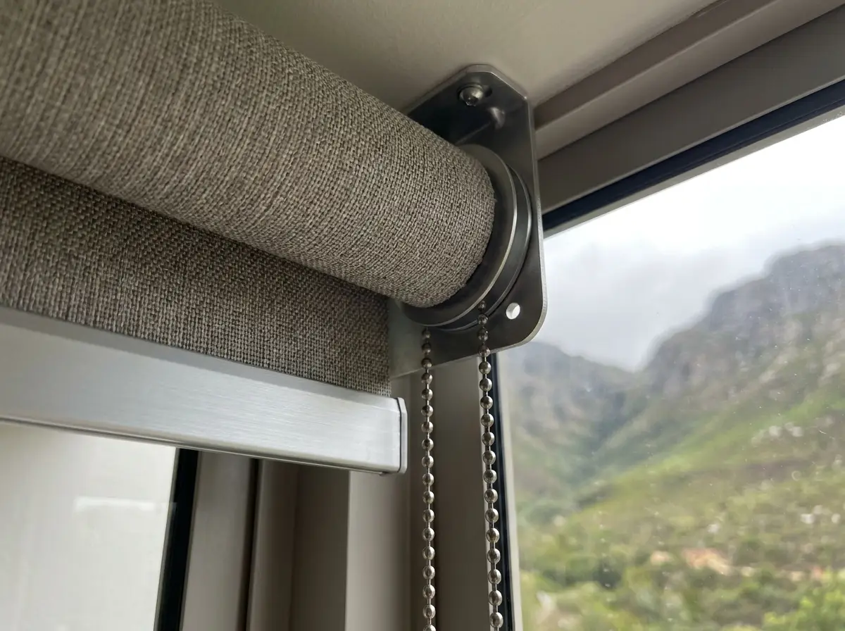 Close-up of a roller blind roller tube and aluminium bracket with pull chains, blurred green kloof mountainside visible through the window behind